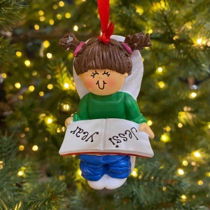 May include: A Christmas ornament featuring a girl sitting on a toilet holding a book that says "Jess! Year". The girl has brown hair, a green shirt, and blue jeans. The ornament is white with a red ribbon.