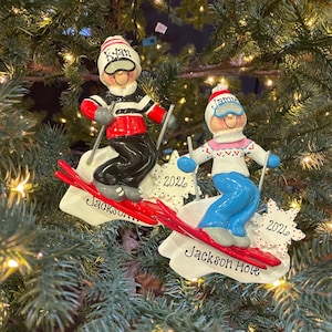May include: Two personalized Christmas ornaments featuring skiers on white bases. One skier wears a red and black outfit with the name Ryan, the other a white and pink sweater with the name Jamie. Both bases are marked Jackson Hole and 2026.