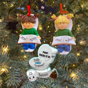 May include: Two hanging ornaments of children with brown and blonde hair wearing green shirts and blue pants, each holding an open book labeled Jessi year and Jayla year. A third ornament shows a toilet with text Poop! There it is! Potty Trained! and a toilet paper roll labeled 2026 Benson.