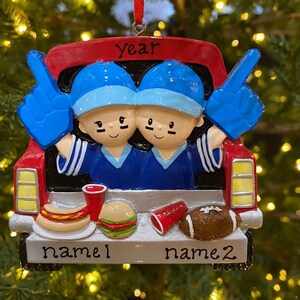 May include: Red truck Christmas ornament with two figures wearing blue and white jerseys and holding up blue number one hands. The truck bed has a hot dog, hamburger, and football. The ornament says "year" and has two blank spaces for names.