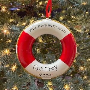 May include: Red and white life preserver Christmas ornament with gold rope trim. The ornament reads "Still Plays With Boats" and "Cpt. Troy - 2023 -"