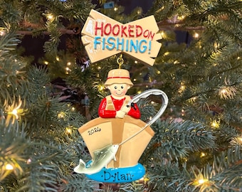 May include: A Christmas tree ornament featuring a person in a tan hat and red shirt sitting in a tan boat. A silver fishing rod extends to a silver fish. A sign above reads HOOKED ON FISHING! The boat displays the year 2026 and the name Dylan.