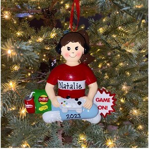 May include: A personalized Christmas ornament featuring a girl wearing headphones and holding a video game controller. The ornament is decorated with a bag of chips, a can of soda, and a "Game On!" sign. The ornament also includes the name "Natalie" and the year "2023".