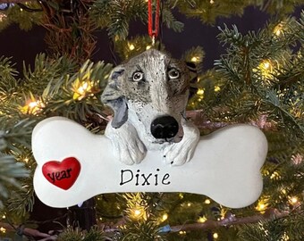 May include: White bone-shaped Christmas ornament with a grey and white dog's head peeking over the top. The ornament has a red heart with the word "year" on it and the name "Dixie" written on the bone.
