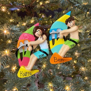 May include: Two colorful personalized Christmas ornaments depicting a boy and girl windsurfing. The boy's ornament has the name "Ben" and the year "2022" on it. The girl's ornament has the name "Katie" and the year "2022" on it.