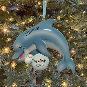 May include: A light blue dolphin Christmas ornament with the name "Hannah" and "Key West 2023" printed on it. The ornament is hanging on a Christmas tree with lights.