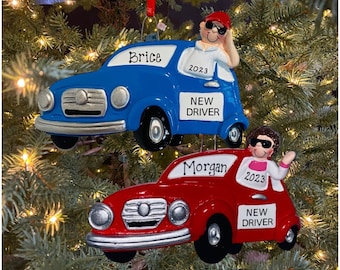 May include: Two Christmas ornaments shaped like cars, one blue and one red. Both cars have a person in the driver's seat, and both have a sign that says "New Driver". The blue car has the name "Brice" and the year "2023" on it. The red car has the name "Morgan" and the year "2023" on it.