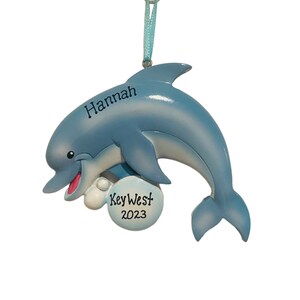 May include: A light blue dolphin ornament with the name "Hannah" and "Key West 2023" printed on it. The dolphin has a smiling face and is holding a white ball. A light blue ribbon is attached for hanging.