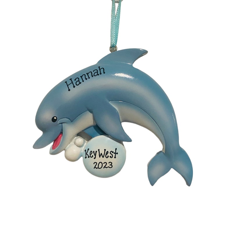 May include: A light blue dolphin ornament with the name "Hannah" and "Key West 2023" printed on it. The dolphin has a smiling face and is holding a white ball. A light blue ribbon is attached for hanging.