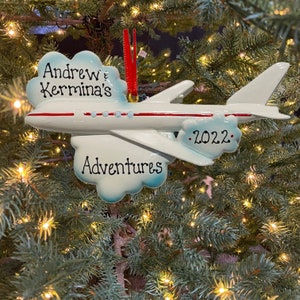 May include: A white airplane Christmas ornament with red accents and the text "Andrew & Kermina's Adventures .2022." The ornament is hanging on a Christmas tree with lights.