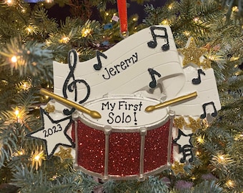 May include: A red and silver glitter drum with the words "My First Solo!" and the year "2023" on it. A white banner with musical notes and the name "Jeremy" is attached to the drum. This is a Christmas ornament.