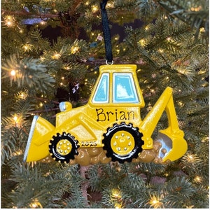 May include: A yellow bulldozer Christmas ornament with black wheels and a black hanging cord. The ornament has the name "Brian" and the year "2023" printed on it. The bulldozer is set against a backdrop of a Christmas tree.
