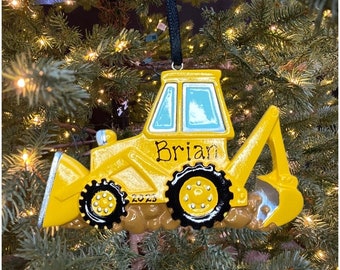 May include: A yellow bulldozer Christmas ornament with black wheels and a black hanging cord. The ornament has the name "Brian" and the year "2023" printed on it. The bulldozer is set against a backdrop of a Christmas tree.