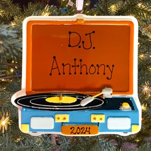 May include: A blue and orange Christmas ornament shaped like a vintage record player with a black record and a needle. The ornament has the text "DJ Anthony 2024" on it.