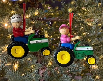 May include: Two personalised Christmas decorations featuring a green tractor with a yellow wheel. The tractor is driven by a person with a red shirt and blue trousers. The ornament says "Sterling" and "2023". The second ornament features a person with a pink shirt and blue trousers. The ornament says "Amelia" and "year".