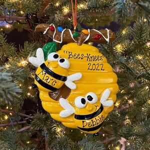 May include: A yellow and black bee-themed Christmas ornament. The ornament features a beehive with two cartoon bees, the names "Marc" and "Debbie", and the text "Bees-Knees 2022". The ornament is hanging on a Christmas tree.