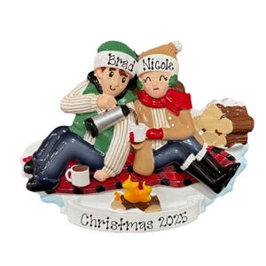 May include: A Christmas ornament depicting a couple enjoying hot drinks by a campfire. The ornament includes the names "Brad" and "Nicole" on their hats, and the text "Christmas 2025" at the base. They are seated on a red and black plaid blanket.