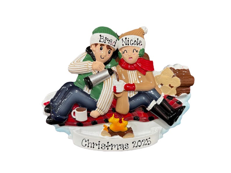 May include: A Christmas ornament depicting a couple enjoying hot drinks by a campfire. The ornament includes the names "Brad" and "Nicole" on their hats, and the text "Christmas 2025" at the base. They are seated on a red and black plaid blanket.