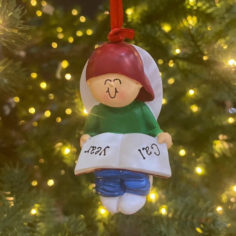 May include: A Christmas ornament featuring a cartoon child wearing a red hat and green shirt, holding a book with the words "Cail" and "Year" written on it.