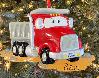 May include: Red and gray dump truck Christmas ornament with the name "Sam" and the year "2023" on the front. The truck is on a brown dirt base with gray rocks.