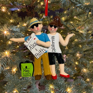May include: A Christmas ornament featuring a couple wearing sunglasses and holding a map. The man is wearing a blue shirt and brown pants, and the woman is wearing a white tank top and black pants. The map says "MAP" and "Alex & Faith". The woman's shirt says "California". A green backpack with the text "May 2023" is in the foreground.