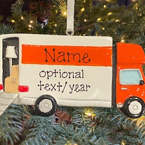 May include: A white and orange moving truck Christmas ornament with the words "Name" and "optional text/year". The truck has a ramp, a lamp, and a box inside. The ornament is hanging on a Christmas tree.