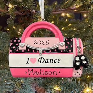 May include: A pink and black dance bag Christmas ornament with the text "2025", "I ♥ Dance", and "Madison". The ornament has a handle and a charm of black ballet shoes. The bag is decorated with polka dots.