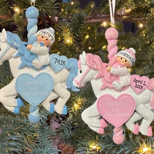 May include: Two Christmas ornaments shaped like carousel horses. One is blue and white with a baby wearing a hat that says "Henry". The other is pink and white with a baby wearing a hat that says "Vivienne Ka". Both have "Baby's 1st Christmas" on a heart.
