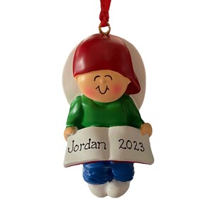 May include: A personalized Christmas ornament featuring a cartoon child wearing a red hat and green shirt, holding a book with the name "Jordan" and the year "2023" written on it.