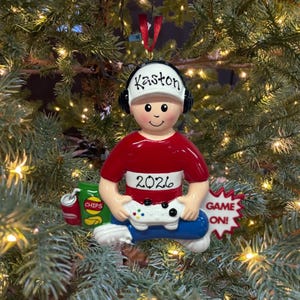 May include: A Christmas tree ornament shaped like a person wearing a white cap with the name Kaston, black headphones, and a red shirt with the year 2026. The figure holds a white game controller, with a bag of chips and a red soda can nearby.