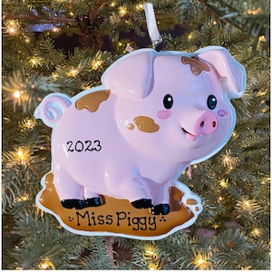 May include: A pink pig ornament with brown mud spots, standing in a brown mud puddle. The ornament has a white ribbon for hanging and says "2023" and "Miss Piggy" on it.