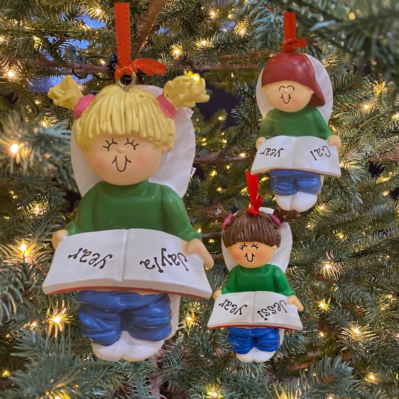 May include: Three Christmas ornaments of children with blonde, brown, and red hair, each wearing a green shirt and blue pants, holding a book with their name and the word "year" on it.
