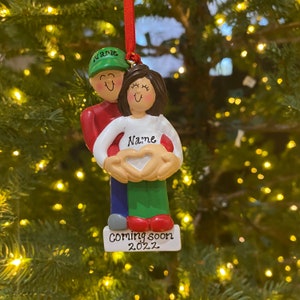 May include: A Christmas ornament depicting a couple expecting a baby. The man is wearing a green hat and a red shirt. The woman is wearing a white shirt with the word "Name" on it. The ornament has a heart shape in the middle and says "Coming soon 2022" at the bottom.