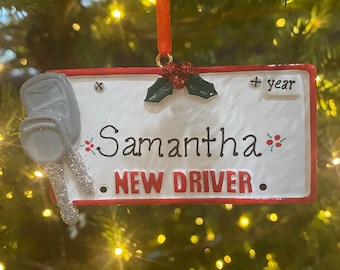 May include: Red and white Christmas ornament shaped like a license plate with the text "Samantha NEW DRIVER" and a silver car key charm. The ornament also has a holly sprig and the text "+ year".