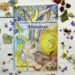 May include: A book cover with a watercolor illustration of a hare, a bird, and a sun with a face. The book title is "Country Wisdom and Folklore Almanac" by Anne Marie Lagram.