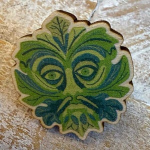 May include: Wooden Green Man face with a leaf-like design. The face is painted with shades of green and blue, with detailed eyes and a beard. The edges of the wood are slightly burnt, giving it a rustic look.