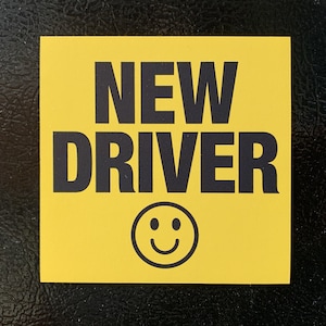 May include: Yellow sign with black text that reads "NEW DRIVER" and a black smiley face.