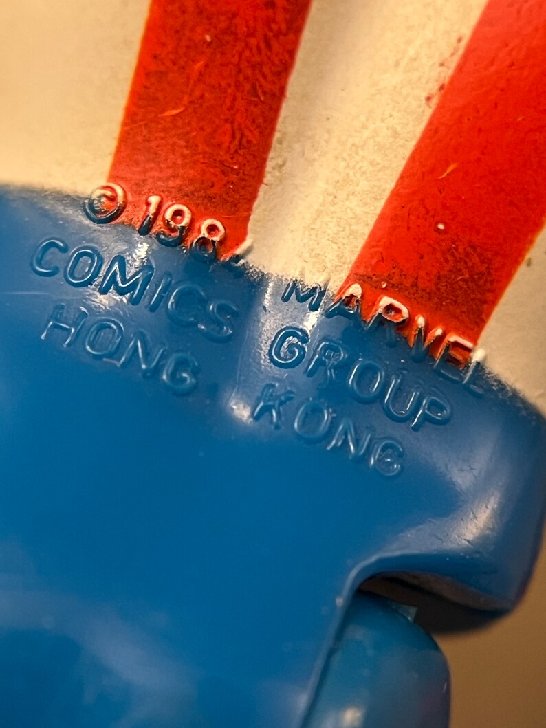 Może przedstawiać: Zbliżenie na niebieską plastikową zabawkę w czerwono-biało-niebieskie paski. Zabawka ma nadrukowany tekst "&copy; 1987 COMICS GROUP HONG KONG" i "MARVEL".