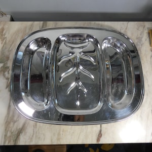 Pode incluir: Uma bandeja de servir prateada com três compartimentos. O compartimento central tem um design decorativo de um galho de árvore. A bandeja é oval e tem um acabamento brilhante.