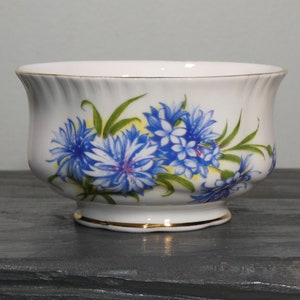 Puede incluir: Un azucarero de porcelana blanca con un diseño floral azul y amarillo. El azucarero tiene un borde festoneado y un borde dorado alrededor de la base.