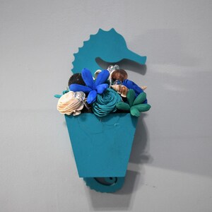 May include: A teal blue ceramic seahorse wall planter with a variety of blue and white flowers and seashells.