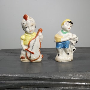 May include: Two vintage porcelain figurines of children playing music. The figurine on the left is a boy wearing a yellow shirt and a red feathered hat, playing a cello. The figurine on the right is a boy wearing a blue shirt and a yellow hat, playing a drum.
