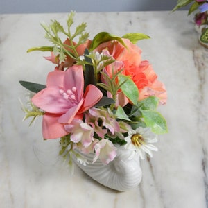 May include: A white ceramic seashell planter with a pink and white floral arrangement. The arrangement includes pink carnations, white bells, and green foliage.