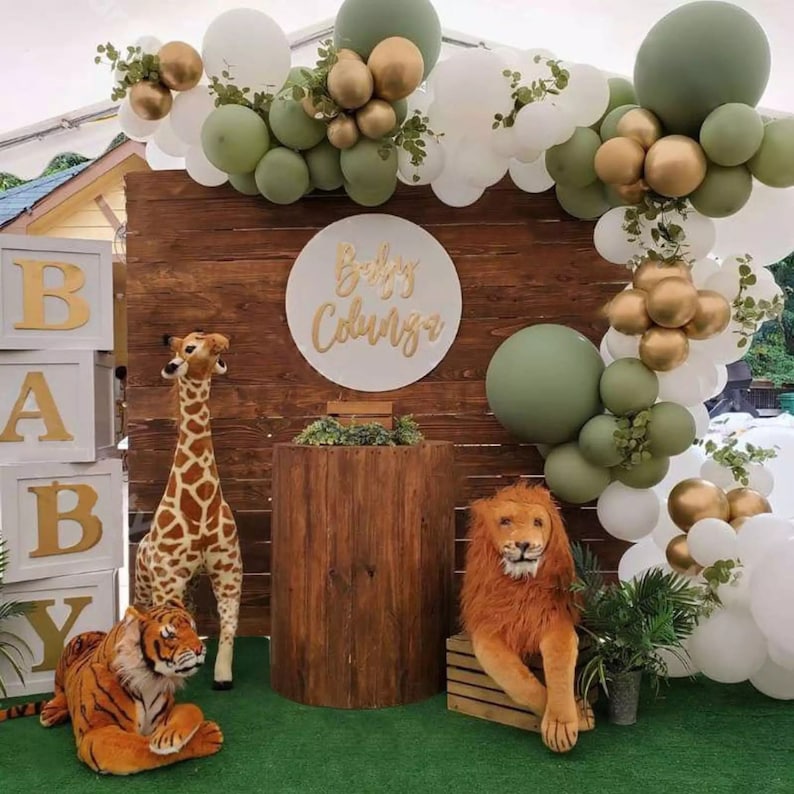 May include: A baby shower scene with a wooden backdrop, featuring a balloon arch in white, gold, and green. Plush animal toys, including a giraffe, tiger, and lion, are placed in front. A sign reads "Baby Colunga".