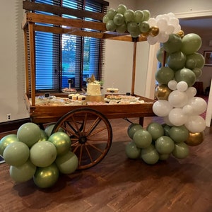 May include: A wooden dessert cart with a variety of treats, including a cake, cupcakes, and cookies. The cart is decorated with green, white, and gold balloons. The cart has a rustic design with a large wooden wheel.