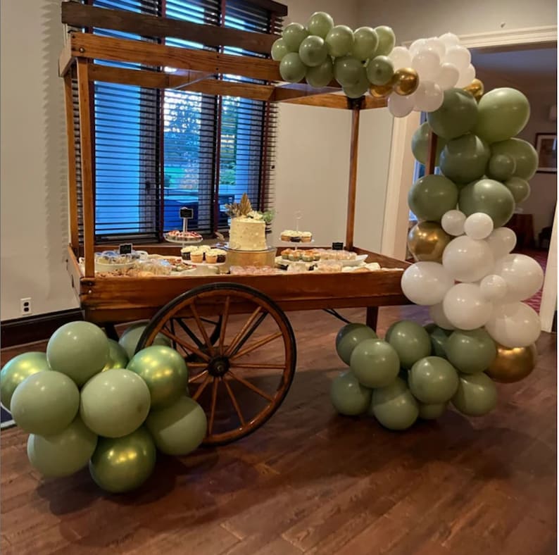 May include: A wooden dessert cart with a variety of treats, including a cake, cupcakes, and cookies. The cart is decorated with green, white, and gold balloons. The cart has a rustic design with a large wooden wheel.