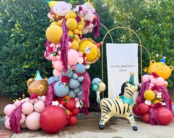 May include: A festive balloon garland display featuring a mix of pink, red, yellow, blue, and brown balloons, accented with artificial flowers and fringe. Includes foil animal balloons, a zebra balloon, and a banner reading HAPPY BIRTHDAY Emma.