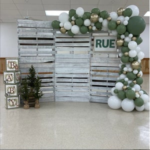 May include: A rustic backdrop for a baby shower or event, featuring a wooden pallet wall, a balloon arch in white, green, and gold, and decorative blocks spelling "BABY". The sign above the backdrop reads "RUE".