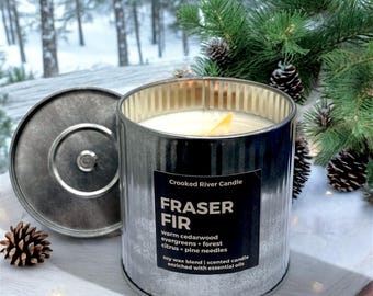 May include: A cylindrical metal candle holder with a lid, containing a Fraser Fir scented candle. The candle has a white wax and a yellow wooden wick. The label reads "FRASER FIR" and lists the scent notes. The background includes pine branches and a snowy forest.