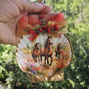 May include: A decorative seashell ornament featuring a printed illustration of three brown horses running through a field of red flowers. The shell is topped with a sheer red ribbon bow, small green sprigs, and red berries, hanging from a red string.